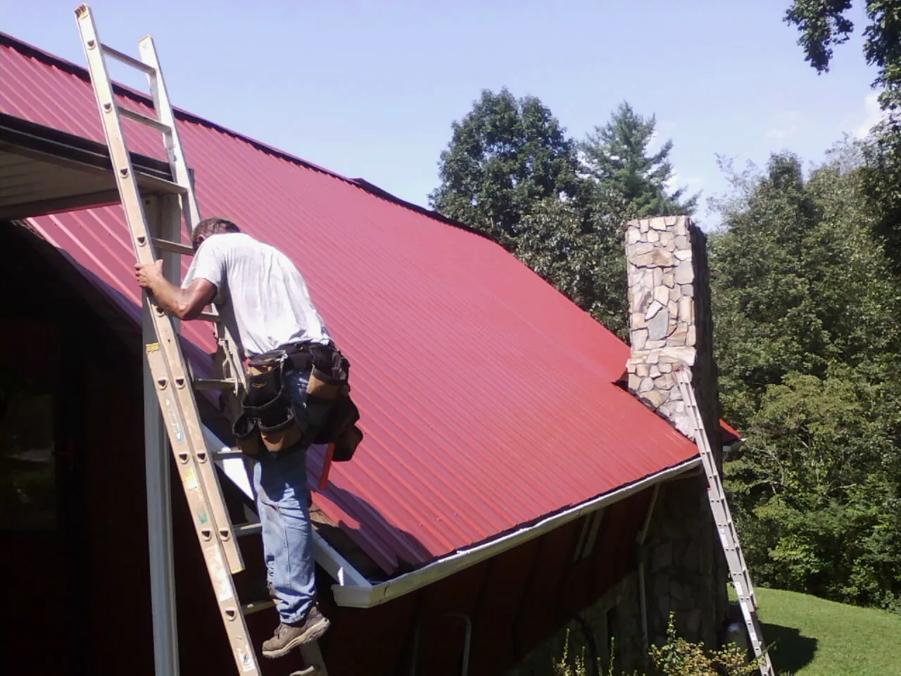 Roofer on a ladder beside a red metal roof and stone chimney — Soffit & Fascia Repair by Roofing Brothers in Saint Helens